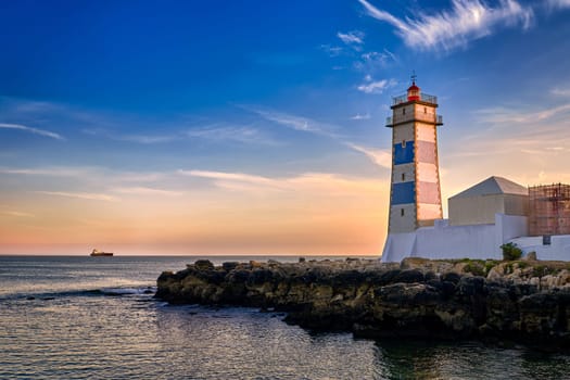 Dramatic sunset on sea shore and Santa Marta lighthouse in Cascais, Portugal. Colorful sky, clouds, sunlight, low sun, calm ocean waters, local landmark, safety and navigation tower, rocky shore.