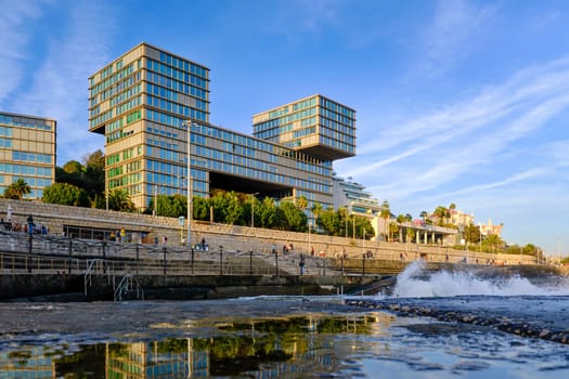 FEB 17, 2024 - Cascais, Portugal: Estoril Sol Residence, luxury block of apartments by waterfront, ocean pool and next to railway to Lisbon on sunny day, tidal waves splash on promenade. Modern architecture by Goncalo Byrne Arquitectos