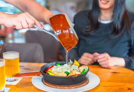 Focus on the hands of a restaurant waiter holding a ceramic frying pan and pouring melted cheese onto a cast iron dish filled with bread croutons and colorful vegetables to a smiling woman customer.
