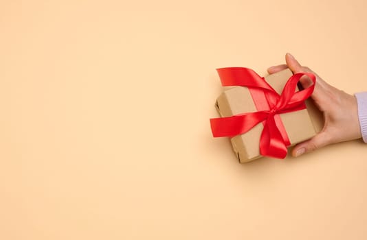 A woman's hand holds a gift box wrapped in a red silk ribbon on a beige background, top view
