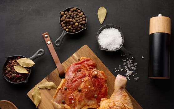 Raw chicken drumsticks seasoned on a wooden board, accompanied by salt and peppercorns, viewed from the top. Black table