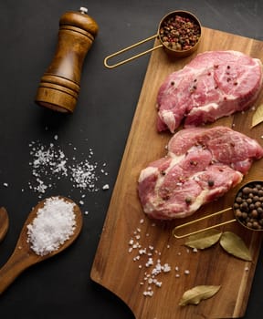 Two raw pork neck steaks on a board and spices for cooking. Top view of black table.