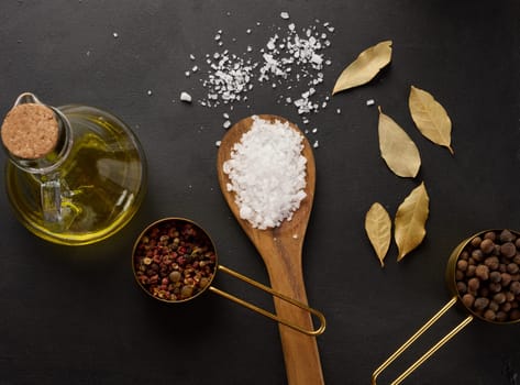 Salt, allspice, peppercorns and olive oil in a bottle on a black table, top view
