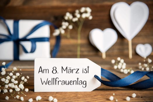 Label With German Text Am 8. Maerz Ist Weltfrauentag Means March, 8th Is International Womens Day. White Festive Decoration Like Present With Blue Bow, Heart and Flowers. Vintage Wooden Background.