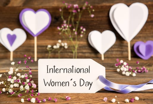 Label With English Text International Womens Day. Purple And Lilac Decoration And Spring Flower Arrangement. Heart Symbols With Wooden Background.