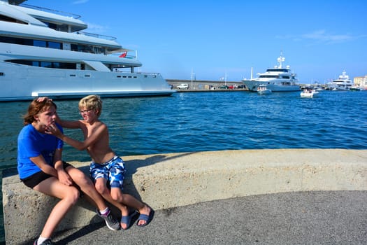 A girl and a boy sit on a wall at the harbor with funny facial expressions, big ships in the background