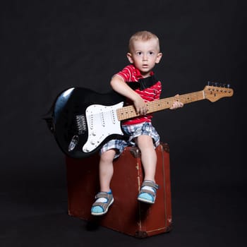 Small boy in striped shirt and checkered shorts sits on an old suitcase with an electric guitar