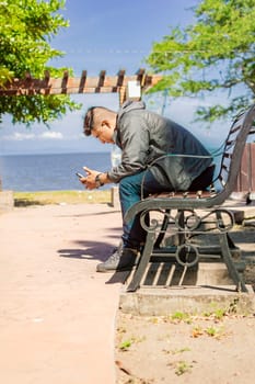 man sitting on a bench checking his phone, young man checking his cell phone sitting on a bench