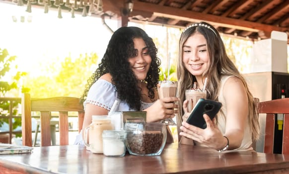 Two teenage girls taking a selfie in a coffee shop, Two happy female friends checking the cell phone in a coffee shop, Teenage girl showing the cell phone to her friend in a coffee shop