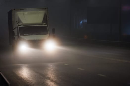 A vehicle with automotive lighting is rolling down the foggy highway at midnight. The headlamps illuminate the asphalt road surface as it travels through the dark night