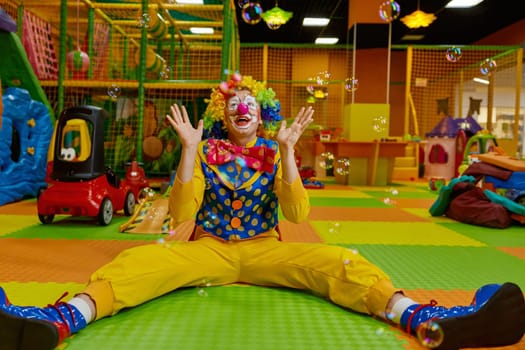 Funny childish clown feeling excitement and happiness playing with flying soap bubbles while sitting at playroom