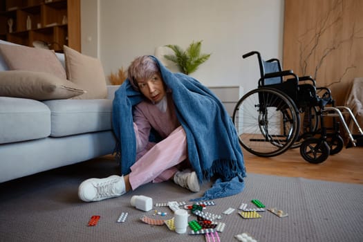 Scared senior woman with mental disorder wrapped in blanket sitting on floor with scattered pills, tablets. Elderly female suffering from panic attack and uncontrollable fear. Loneliness and sickness
