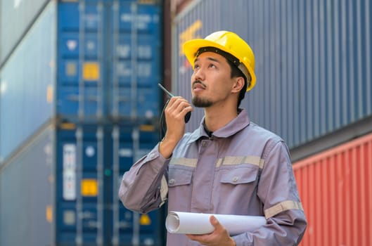 Japanese male engineer or foreman in uniform with yellow hardhat using walkie talkie to control loading containers at cargo shipping yard. Transportation and shipping concept
