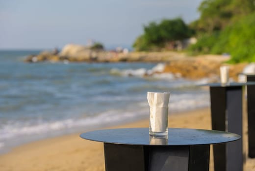 Glass of water with white napkin on a wooden table on a sandy beach. The ocean is visible in the background, with gentle waves lapping at the shore.