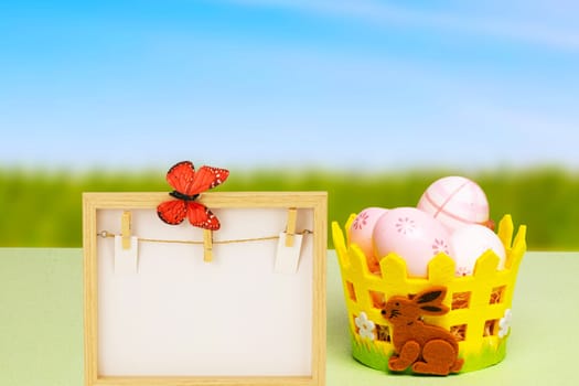 Easter greeting card template. Colorful Easter eggs in a basket and an empty wooden frame with a butterfly on table over natural blurred sky background.