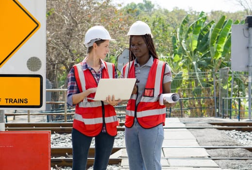 Two individual engaged in a discussion at a railway site, wearing safety equipment, including hard hats and high-visibility vests holding a laptop computer and rolled-up document.