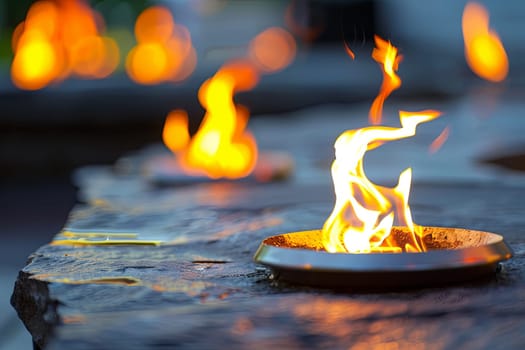 A close-up view of a metal fire pit filled with burning logs and bright orange flames dancing upwards. The fire pit is surrounded by stones and embers, creating a warm and glowing atmosphere.