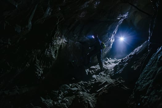 A man standing in a dark cave shines a flashlight to illuminate the surroundings. The cave walls are rugged and uneven, with stalactites hanging from the ceiling. The man appears focused and determined as he explores the cave.