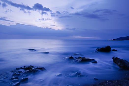 Waves crash against rocks in the foreground as the ocean stretches into the distance, captured in a long exposure shot.