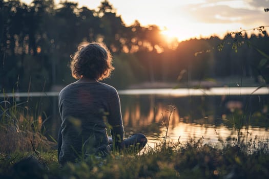 A person is seated on a grass field next to a body of water. The individual appears relaxed and is enjoying the natural setting surrounding them.