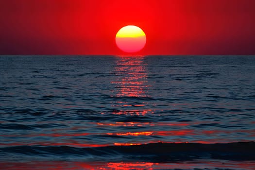 In the scene, the sun sets over the ocean, casting a vivid red and yellow glow across the sky and water. The horizon is illuminated with warm hues as the sun dips below, creating a stunning display of colors.