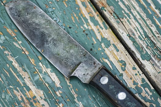 A large knife is placed on top of a rustic wooden table, creating a stark and striking visual contrast between the sharp metallic blade and the natural wood surface. The knifes handle is textured and the blade appears sharp and well-crafted.