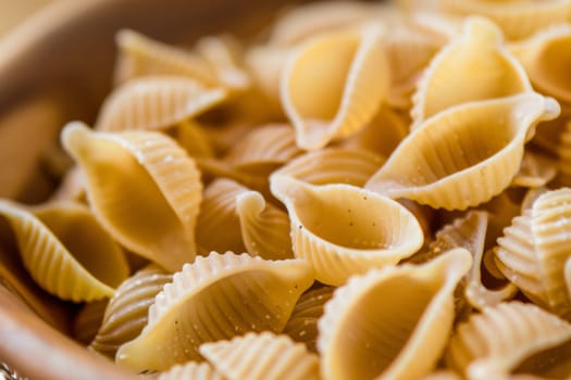 A wooden bowl filled with pasta shells sits atop a table, showcasing the variety of shapes and textures of the pasta. The warm wooden tones of the bowl contrast with the table surface, creating a visually appealing scene.