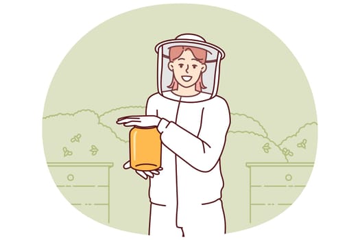Young woman beekeeper in protective uniform holding jar of honey standing in apiary among flying bees. Girl farmer breeds bees in garden getting organic honey for sale at fair of ecological products