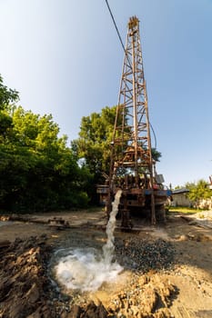Old water pump truck extracts water from a hole in the ground at sunny summer day, surrounded by a natural landscape with trees, plants, and soil. Ultra-wide angle.