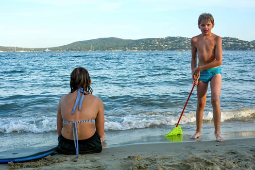 A girl sits by the sea in front of the waves, a boy with a fish net stands on the sandy beach