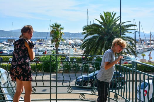 Two children stand on a balcony with an astonished expression on their faces, in the background the harbor with many boats, street and palm tree