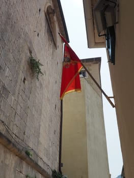 The national flag of Montenegro has a red field with gold border and coat of arms of Montenegro in its center. Symbol of the state of Montenegro. Light breeze. The flag is hung on street of old city