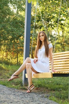 Young woman in white sundress sitting on swing in city park