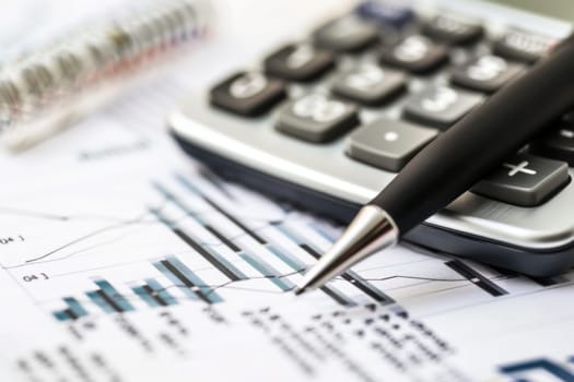 A black pen is positioned on top of a silver calculator, which in turn rests on a stack of various papers. The pen is still and the calculator displays numbers, suggesting work in progress.
