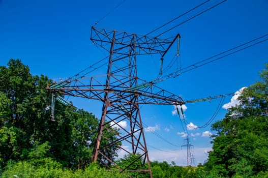 The towers of the power line of energy with cables against the background of nature. High -voltage tower of transmission of electric energy. Industry and nature. Blue sky with white clouds.