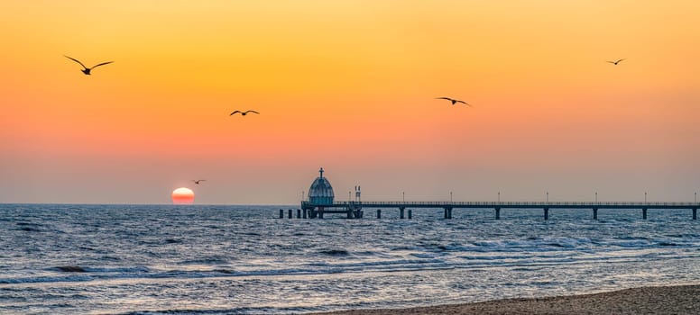 Orange sunrise over the Zinnowitz pier on Usedom, with seagulls flying over the sea.
