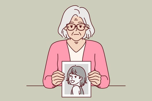 Elderly woman remembers youth, showing portrait from past, and looks at screen with slight sadness. Elderly lady with gray hair shows photo of herself from childhood, wishing she could turn back time.