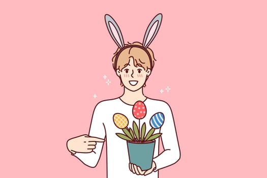 Guy celebrating easter holds pot of plants in shape of multi-colored eggs, wears bunny ears on head
