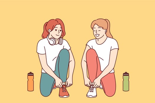 Two women runners prepare for training, tying shoelaces and discussing details of marathon or sprint competition. Female runners use wireless headphones to listen to music and podcast while training