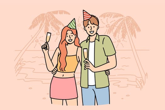 Honeymoon happy couple drinking champagne, standing on beach of sunny resort, wearing birthday caps. Romantic guy and girl celebrate beginning of honeymoon on ocean shore with palm trees