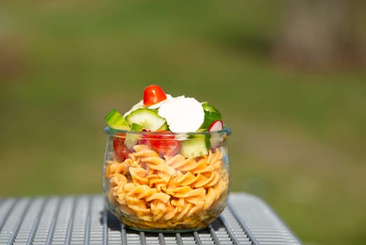 Pasta salad in a bowl, sliced cucumber, tomato, red pepper and cheese, healthy food with vegetables, picnic in nature