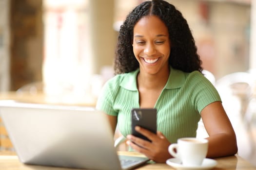 Happy black woman using phone and laptop in a bar terrace