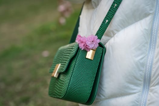 Women's green crossbody bag with pink japanese cherry blossoms on the shoulder of woman in white jacket while walking in spring park.
