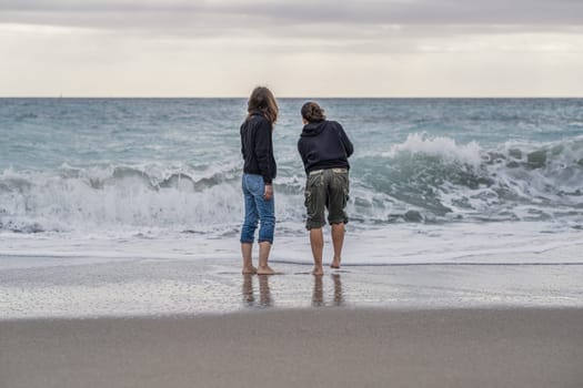 Mother and daughter barefoot on the shoreline, looking at something on the horizon, rear view A young mother spending time on the beach with her teenage daughter, conceptual theme concerning family and femininity.