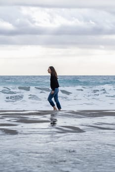 Adult woman with long brown hair walks alone and carefree on the shoreline barefoot. Full-length side view, casual wear with jeans and winter jumper. Vertical composition, copy space.