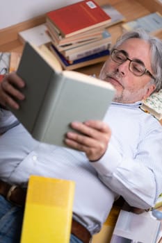 A man with a serene expression lying on the wooden floor intent on reading a book. He is wearing a shirt and reading glasses and is surrounded by many books. Concept of leisure, education and passion for reading.