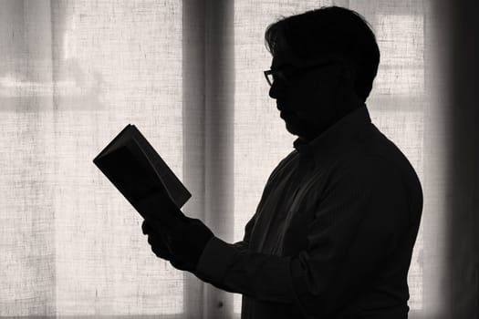 Profile view of a mature man focused on reading a book, male silhouette standing in front of a window with white curtain. Relaxing and instructive leisure concept.