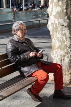 Mature adult sitting on a bench using his phone. Man with white hair and beard dressed in a leather jacket and rust pants busy in his phone. Outdoor. City lifestyle.