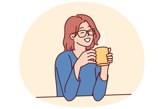 Joyful woman holding coffee mug sitting at table and smiling remembering happy moments from life during lunch break. Girl in casual clothes drinks hot tea or coffee to gain vigor and energy