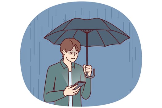 Sad man with umbrella stands in rain and reads SMS in mobile phone from girlfriend who refused to come on date. Concept of autumn depression due to bad weather and weather sensitivity